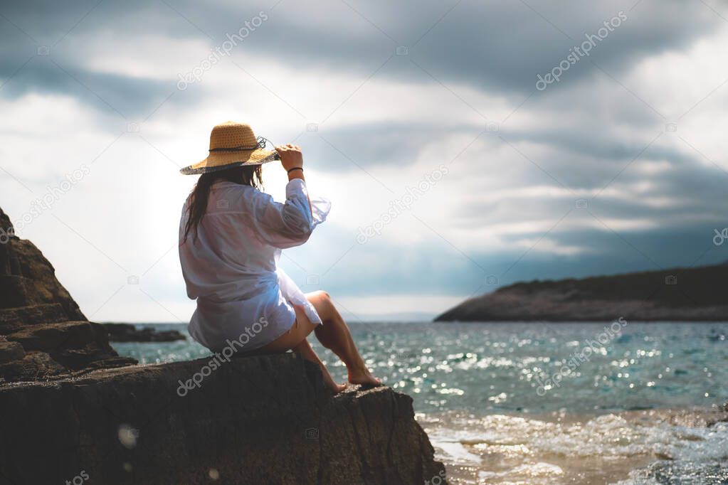 Beautiful brunette seen from behind sitting on a rock on the shores of adriatic sea, cloudy day during the summer of 2021. Waves hitting the shore