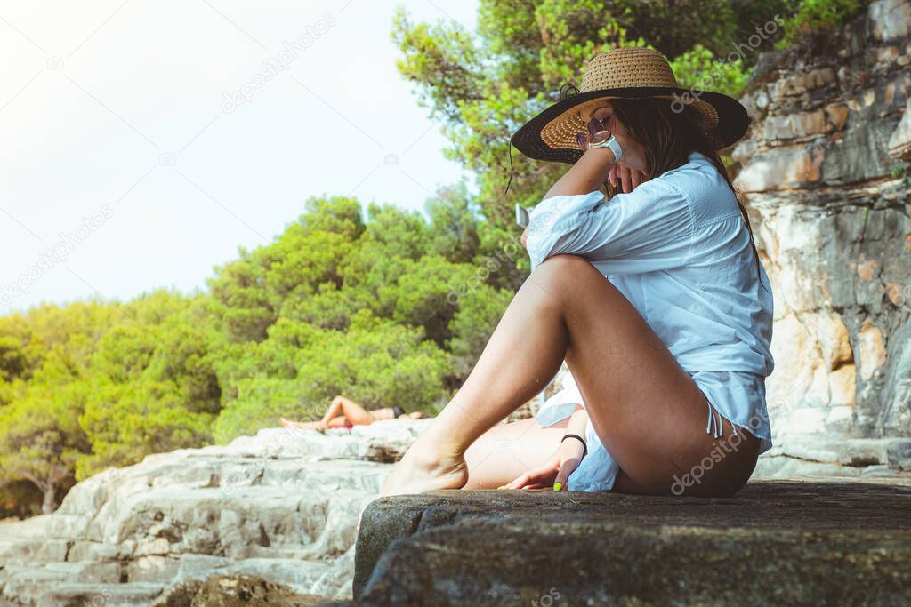 Beautiful brunette sitting on a rock on a beach in Croatia. Wearing a hay hat and a white shirt, warm sunny day, happy smile while posing outside.