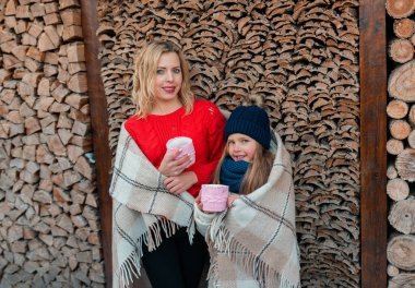 Çay termosuyla gülümseyen sevimli bir aile, battaniye ile kaplı, arka planda yakacak odunlarının yanında oturuyor. Kavram, soğuk, kış, sıcak giysi konsepti