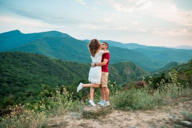 Genç çift dağlarda el ele tutuşup birbirlerine bakarak yürüyor.