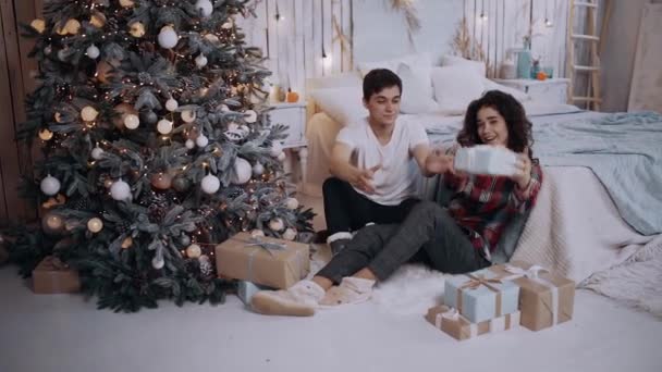 Pár, fiú és lány, leülnek a nappaliban a karácsonyfa mellett.. — Stock videók