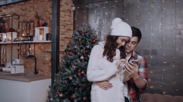 Junges Paar umarmt sich zu Hause am Weihnachtsbaum und verfolgt mit seinem Handy die Rabatte für das neue Jahr. — Stockvideo