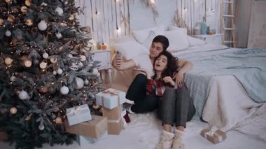 Genç çift evde Noel ağacının yanında otururken telefonla sarılıp fotoğraf çekiyorlar..