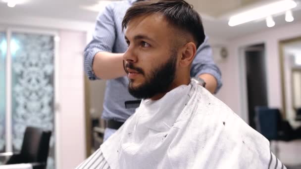 Jongeman in de kapperszaak zit in een fauteuil. Kapper knipt zijn haar met een trimer. — Stockvideo