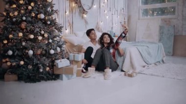 Genç çift evde Noel ağacının yanında otururken telefonla sarılıp fotoğraf çekiyorlar..