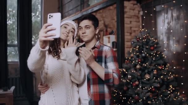 Young couple dressed in Christmas clothes, having fun and taking pictures with their phone near the Christmas tree — Stock Video
