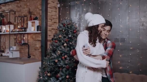 Pareja joven abrazándose en casa cerca del árbol de Navidad, usando su teléfono móvil para rastrear los descuentos de Año Nuevo — Vídeos de Stock