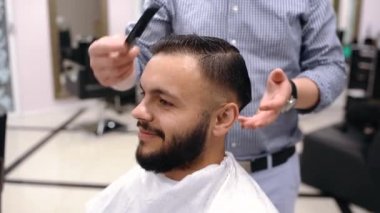 Profesyonel erkek kuaför genç pozitif bir adamın saçını makasla keser, kameraya bakar ve gülümser.