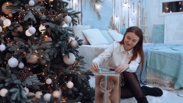 Mujer caucásica en traje acogedor envuelve una caja de regalo en casa decorada de Navidad — Vídeo de stock