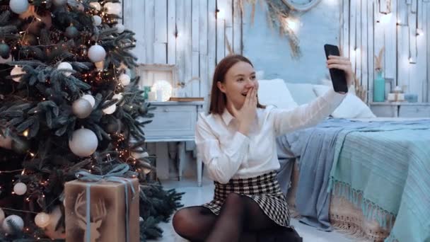 Az izgatott fiatal nő telefonon beszél a családjával, a karácsonyfa mellett ülve., — Stock videók