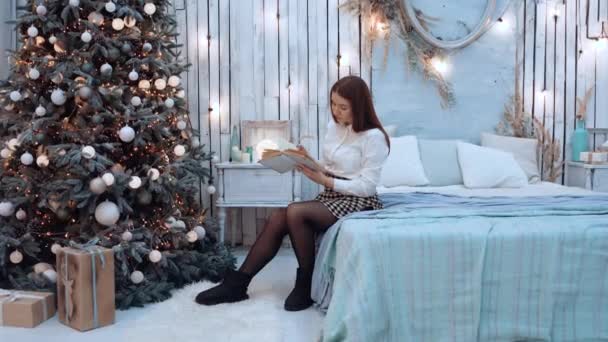 Kvinna tillbringar fritid med att läsa en bok nära den dekorerade julgranen. — Stockvideo
