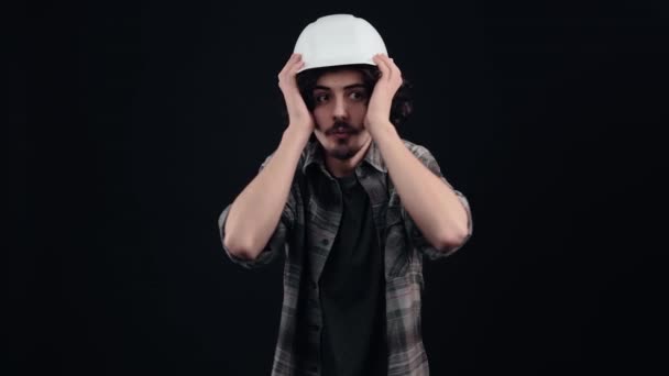 Ein überraschter Ingenieur trägt im Atelier einen speziellen Helm auf dem Kopf, bekleidet mit einem karierten Hemd auf schwarzem Hintergrund, — Stockvideo