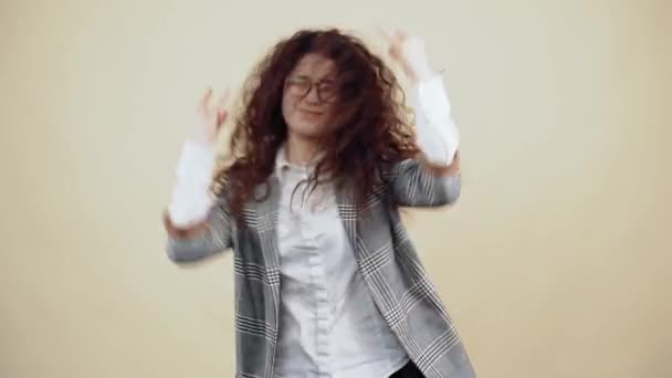 La jeune femme folle danse rock avec des cheveux bouclés dans une veste grise et chemise blanche, avec des lunettes posant isolées sur un fond beige dans le studio. — Video