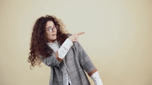 Oye tú, mira allí. Hermosa joven con pelo rizado en chaqueta gris y camisa blanca, con gafas posando aisladas sobre fondo beige en el estudio. El concepto de estilo de vida de las personas. — Vídeo de stock