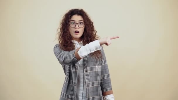 회색 재킷 과 흰색 셔츠를 입은 곱슬곱슬 한 머리에 충격을 받은 젊은 여자, 스튜디오의 베이지 배경에 따로 놓여 있는 안경. — 비디오