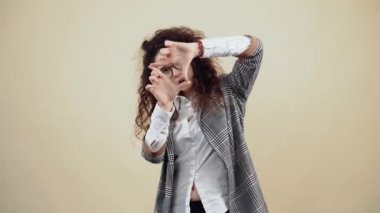 Meraklı genç bayan yeni şeyler bulmak için parmaklarının arasından bakıyor. Gri ceketli, beyaz gömlekli, gözlüklü, stüdyoda bej bir arkaplanda izole edilmiş bir poz veriyor..