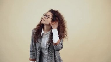 Eli kulağında olan meraklı genç kadın ofis arkadaşlarını dinliyor ve sesler duyuyor. Gri ceketli, beyaz gömlekli, gözlüklü, bej arkaplanda izole bir poz veren...