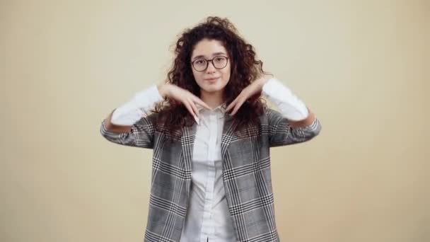 The happy young woman with a wide smile extends both hands in front to offer hugs. Cretaceous in gray jacket and white shirt, with glasses posing isolated on a beige background in the studio. — Stock Video