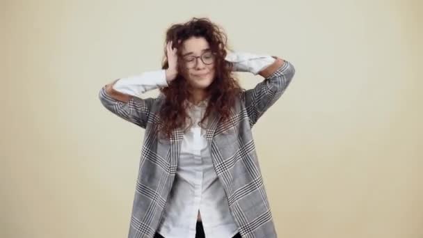 Mujer joven aburrida, enferma, con la mano en el oído para no oír, irritada por el fuerte ruido. Cretácico en chaqueta gris y camisa blanca, con gafas posando aisladas sobre un fondo beige en el — Vídeo de stock