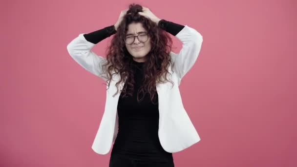 La giovane donna triste ha mal di testa, mettendo le mani sulla testa per eliminare il dolore. Giovane hipster in giacca bianca e camicia nera, con occhiali in posa isolata su sfondo rosa in studio. — Video Stock