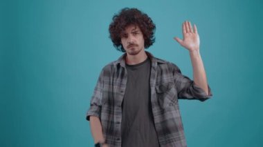 Eli ile selam veren kıvırcık genç adam odaya bakıyor, sonunda gülümsüyor. Ekose gömlek ve koyu renk tişört giyen hippi.