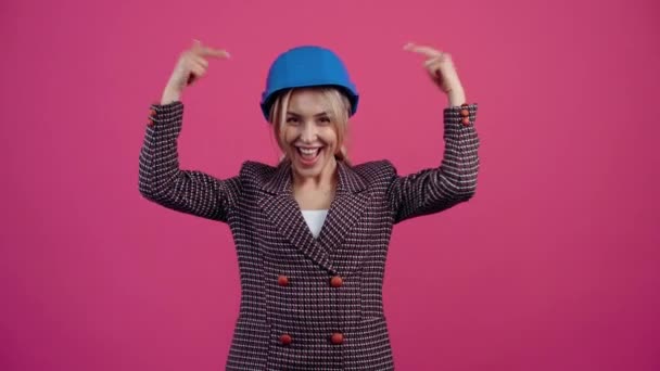 Зрелая блондинка с шлемом инженера на голове синего воротничка, широко улыбается, глядя в комнату молодая зрелая блондинка в розовой куртке. — стоковое видео