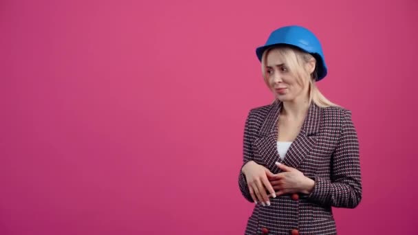 Schöne blonde reife Frau, empfiehlt ein Produkt auf der linken Seite, trägt einen Ingenieurhelm auf dem Kopf Junge reife Blondine in rosa Jacke. Vereinzelt auf rosa Hintergrund. — Stockvideo