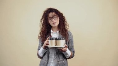 Elinde 4 kahveyle bir destek tutan tebeşir gibi, kibirli sekreter, onları önde sunuyor. Gri ceketli, beyaz gömlekli, gözlüklü, bej arka planda yalnız poz veren genç hippi.