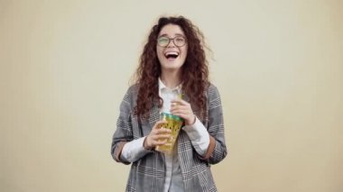 Kıvırcık saçlı genç kadın elinde taze meyve suyu ile sarı bir bardak tutarken yüksek sesle gülüyor. Gri ceketli, beyaz gömlekli, gözlüklü genç hippi bej üzerine izole edilmiş poz veriyor.