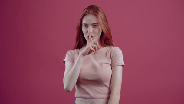 Tânăra roșcată timidă, cu degetul pe buze, arată semnul că tace, promite un secret. 20 de ani, adolescent pistruiat hipster într-un tricou casual roz, izolat pe un fundal roz . — Videoclip de stoc