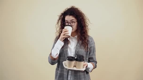 De rijpe Kretenzer houdt in zijn hand een steun met 4 koffies. Hij drinkt uit een glas en wordt wakker. Jonge hipster in grijs jasje en wit shirt, met bril poseren geïsoleerd op beige achtergrond — Stockvideo