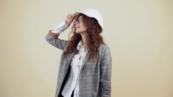 Młoda kobieta z kręconymi włosami nosi biały kask inżynierów, rozgląda się, uśmiecha i pokazuje swój język. Młody hipster w szarej kurtce i białej koszuli, w okularach pozujących na beżu — Wideo stockowe