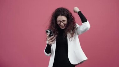Kıvırcık saçlı mutlu genç bir kadın cep telefonunu elinde tutuyor ve spreyine bakıp gülümsüyor. Beyaz ceketli ve siyah gömlekli genç hippi.,