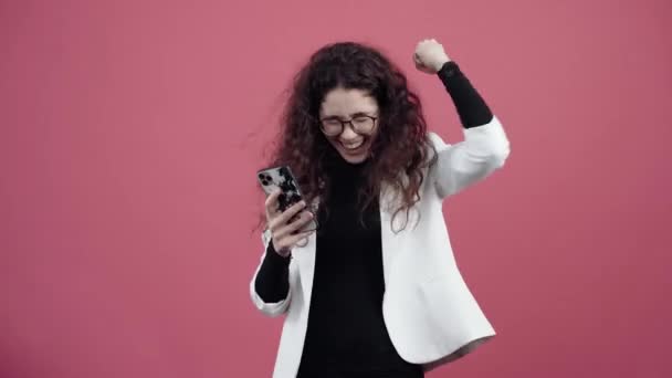 Gelukkige jonge vrouw met krullend haar, houdt haar mobiele telefoon in haar hand, en kijkt naar de spray, glimlachend. Jonge hipster in wit jasje en zwart shirt, — Stockvideo