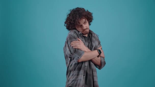 Joven tipo hipster empático. Tímido abraza con sus manos mirando perplejo a la derecha, finalmente sonriendo. Niño vestido con camisa a cuadros gris, sobre fondo estudio azul — Vídeos de Stock