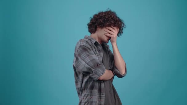 Shy, sad teenager with curly hair. With his hands over his eyes, he spies between his fingers, looking down, and finally looks into the room. Boy dressed in gray plaid shirt, on blue studio background — Stock Video