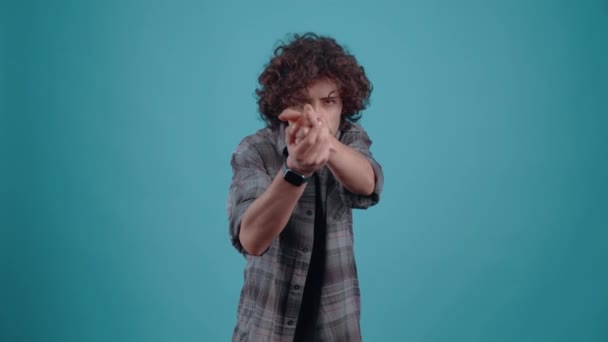 Charismatický hipster napodobuje pistoli rukama a střílí vepředu, oblečený v kostkované košili, s kudrnatými vlasy, ve studiu na tyrkysovém pozadí. — Stock video