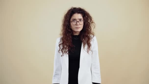 Jovem mulher positiva com cabelo encaracolado aponta seus dedos para ela como um sinal para se inscrever com um sorriso. Jovem hipster em jaqueta cinza e camisa branca, com óculos posando isolado no fundo bege em — Vídeo de Stock