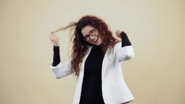 The young woman with curly hair dances with her hands, holds her fingers in her fists, shakes her hands to the left and to the right, fooling around. Young hipster in gray jacket and white shirt, — Stock Video