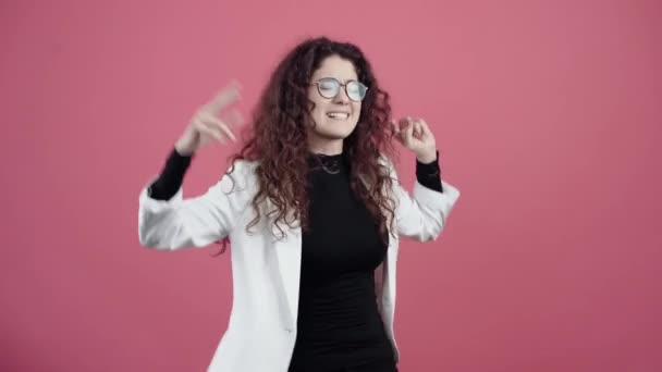 Счастливая молодая женщина с вьющимися волосами танцует руками, два открытых пальца трепещут. Молодой хипстер в белой куртке и черной рубашке, с очками, позирующими изолированно на розовом фоне в студии. — стоковое видео