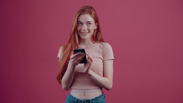 La joven con el pelo rojo con el teléfono en la mano piensa, luego escribe un mensaje y sonríe felizmente. Adolescente hipster pecoso de 20 años con una camiseta casual rosa, aislada sobre un fondo rosa. — Vídeo de stock