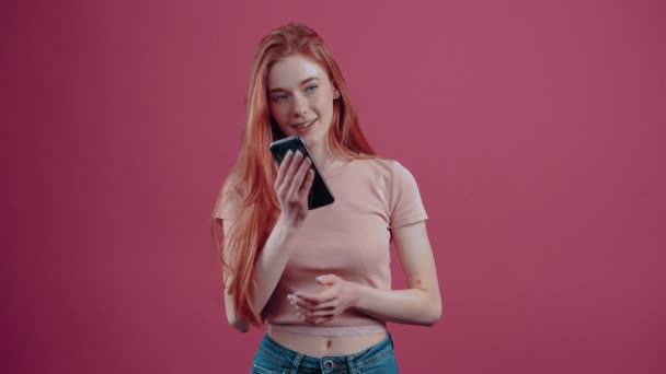 Glad rödhårig ung kvinna pratar i telefon, upphetsad med munnen mot munnen. 20-årig fräknig hipster tonåring i en rosa casual T-shirt, isolerad på en rosa bakgrund. — Stockvideo