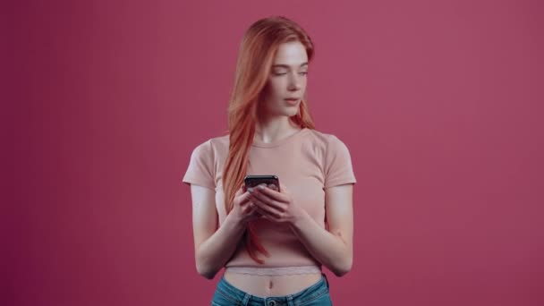 カリスマ的な若い女性は、彼女の手の中に携帯電話を保持しながら、驚いて、右に見て、幅笑顔をお勧めします。ピンクのカジュアルなTシャツに身を包んだ20歳のハイパーティーンアー — ストック動画