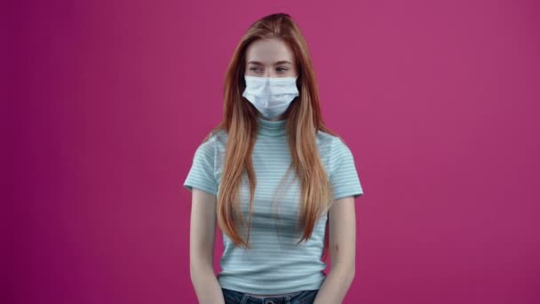 Δυσαρεστημένη κοκκινομάλλα με χειρουργική μάσκα στο πρόσωπό της, αρνείται να κρατήσει τους αντίχειρές της κάτω. 20χρονος φακίδες hipster έφηβος σε ένα ροζ casual T-shirt, απομονώνονται σε ένα ροζ φόντο. — Αρχείο Βίντεο