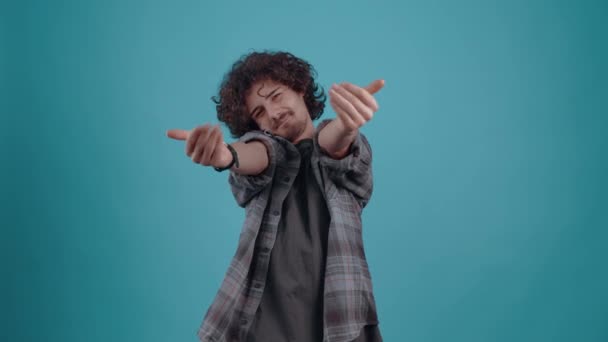 Lindo y emocionado joven hipster, con sus manos quiere abrazar sonriendo. Niño vestido con camisa a cuadros gris, sobre fondo de estudio azul El concepto de estilo de vida de las personas — Vídeos de Stock