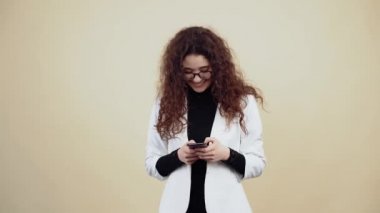 Telefonla mesaj yazan kıvırcık saçlı genç bir kadının geniş gülümsemesi, sonunda kameraya geniş bir gülümseme. Gri ceketli, beyaz gömlekli, gözlüklü genç hippi bej üzerine izole edilmiş poz veriyor.