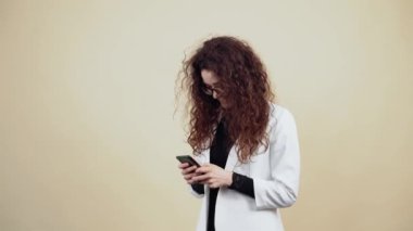 Kıvırcık saçlı mutlu genç kadın elinde bir telefon tutuyor ve mesajla arkadaşlarıyla takılıyor. Gri ceketli, beyaz gömlekli, gözlüklü, bej arka planda yalnız poz veren genç hippi.
