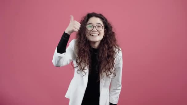 Mujer joven feliz con el pelo rizado, con las manos muestra su pulgar hacia arriba como una señal, recomendando sonreír. Joven hipster en chaqueta blanca y camisa negra, con gafas posando aisladas sobre fondo rosa en — Vídeos de Stock