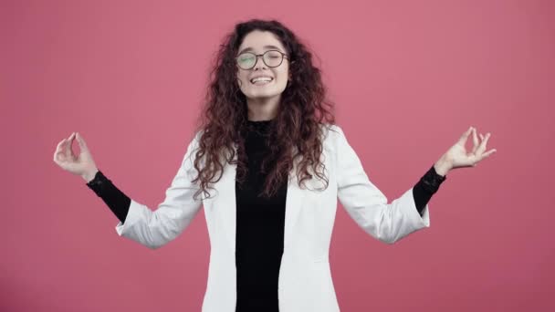 Calm and balanced young woman with curly hair, is inspired by holding her hands in the shape of yoga, relaxing. Young hipster in white jacket and black shirt, with glasses posing isolated on pink — Stock Video