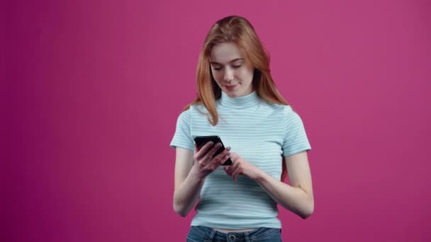 La joven pecosa feliz recibe una llamada y habla con entusiasmo por teléfono, en una camiseta casual rosa, aislada sobre un fondo rosa. El concepto de estilo de vida de las personas — Vídeos de Stock